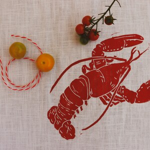 May include: A red lobster print on a white linen tea towel with two small yellow tomatoes and a sprig of red tomatoes.