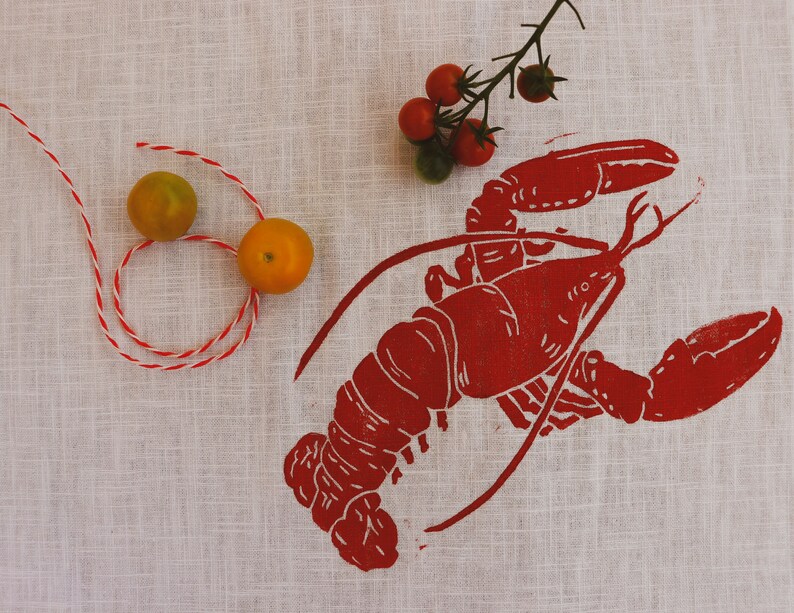 May include: A red lobster print on a white linen tea towel with two small yellow tomatoes and a sprig of red tomatoes.