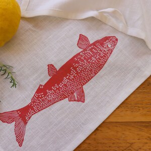 May include: A white linen tea towel with a red fish design printed on it. The fish is facing right and has a detailed design. The towel is lying on a wooden surface.