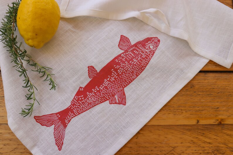 May include: A white linen tea towel with a red fish design printed on it. The fish is facing right and has a detailed design. The towel is lying on a wooden surface.