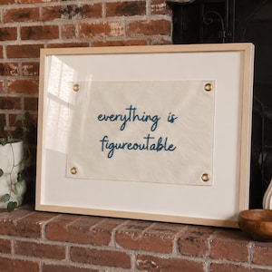 Puede incluir: Un tapiz de pared enmarcado que presenta una bandera de tela color crema con el texto azul oscuro bordado "everything is figureoutable". La bandera está asegurada con cuatro ojales metálicos de tono latón en las esquinas y colocada en un marco de madera clara.