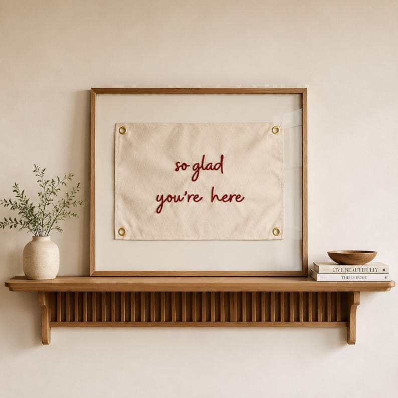 Może przedstawiać: Oprawiony prostokątny baner z pł&oacute;tna z wyhaftowanym ciemnoczerwonym napisem "so glad you're here" oraz czterema złotymi metalowymi oczkami w rogach, wyeksponowany na drewnianej p&oacute;łce obok ceramicznego wazonu z zielenią i książkami.