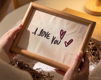 May include: A rectangular wooden frame containing a light-colored fabric panel. The fabric features the embroidered text "I love you" in dark thread, accompanied by two small, stitched heart outlines in a deep reddish-maroon color.