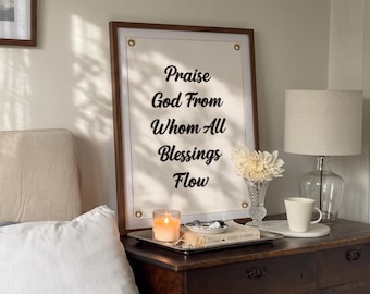 May include: A rectangular wall hanging in a dark wood frame featuring a cream-colored fabric panel with the black text "Praise God From Whom All Blessings Flow" and four brass grommets at the corners, displayed on a dark wooden dresser.