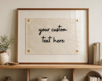 May include: A rectangular wooden frame containing a beige fabric panel with four brass grommets in the corners. The fabric features the black embroidered text your custom text here centered on the surface, displayed within a glass-fronted frame.