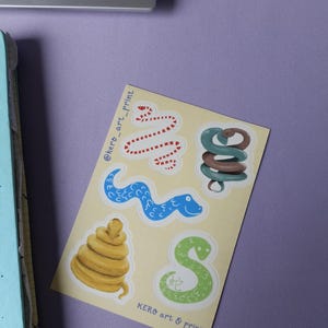 Puede incluir: Una hoja de pegatinas con cinco diseños diferentes de serpientes. Las serpientes están ilustradas en un estilo de dibujos animados con colores brillantes. Las pegatinas están sobre un fondo amarillo con el texto "@kero_art_print" y "KERO art & print" en negro.