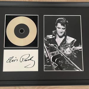 May include: A framed display featuring a gold-colored vinyl record, a black and white photograph of Elvis Presley playing a guitar, and a white rectangular card with the printed signature Elvis Presley in black ink, all set against a black mat.