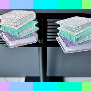 Könnte beinhalten: Zwei dekorative Stapel glitzernder Bücher. Jeder Stapel besteht aus drei Büchern in Weiß, Türkis und Lila. Sie befinden sich auf einer schwarzen, geschlitzten Oberfläche mit grauem Hintergrund.