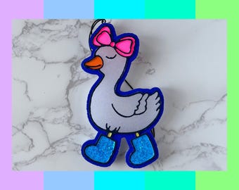 May include: A whimsical, cartoon-style goose ornament with a pink bow, orange beak, and blue boots. The goose is outlined in blue and has a white body. The background is a marble pattern with colorful blocks.