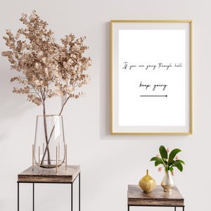 May include: A gold framed print with the text "If you are going through hell, keep going" with an arrow pointing to the right. The print is hanging on a white wall with a vase of dried branches and a small gold vase with flowers on a table.