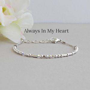 May include: A silver-toned beaded bracelet featuring small metallic spheres and cylindrical spacers with iridescent accents. The bracelet includes a lobster claw clasp and an adjustable chain. The text Always In My Heart is visible.
