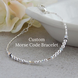 May include: A silver bracelet with small silver beads and clear crystal beads. The bracelet has a clasp and is labeled "Custom Morse Code Bracelet".
