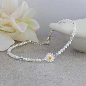 May include: A delicate white beaded bracelet with a daisy flower charm. The bracelet features small white beads, silver accents, and a daisy charm with a yellow center. The bracelet is approximately 7 inches long.
