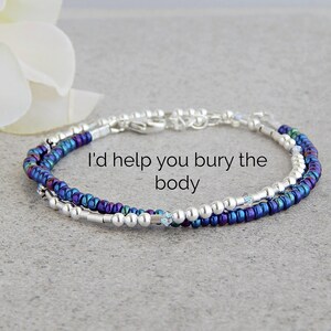 May include: A silver and iridescent blue beaded bracelet with a clasp closure. The bracelet has the text "I'd help you bury the body" on it.