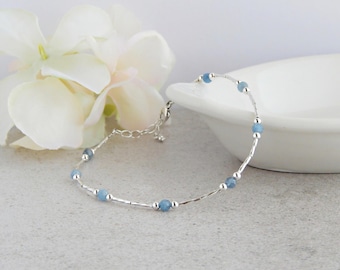 May include: A delicate bracelet featuring small, round, light blue beads interspersed with thin, polished silver-tone metal bars and tiny silver-tone spheres. The jewellery piece includes a silver-tone chain extender with a small clasp closure.