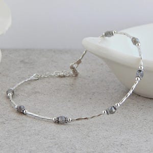 May include: A delicate silver bracelet featuring small, faceted gray beads interspersed with slender, textured silver bars. The bracelet has a simple chain closure and is displayed on a light gray surface.