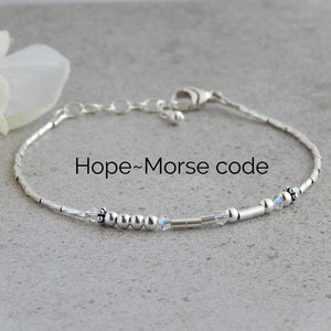 May include: Silver bracelet with small beads and a crystal charm. The bracelet spells out "Hope" in Morse code.
