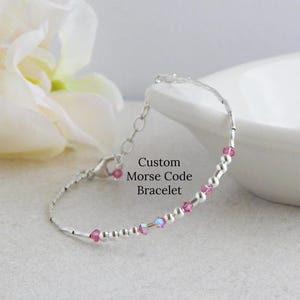 May include: A delicate silver-toned bracelet featuring a sequence of small metallic beads and translucent pink crystal beads arranged in a Morse code pattern. The bracelet includes a lobster claw clasp and a short extender chain. Text reads Custom Morse Code Bracelet.