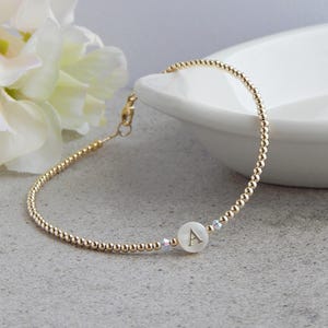 Puede incluir: Un brazalete de cuentas doradas con una sola cuenta blanca con la letra 'A' en oro.