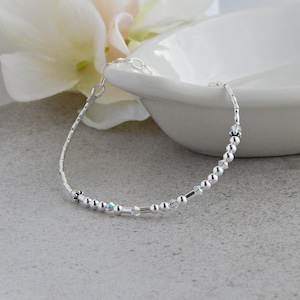 May include: A delicate silver bracelet featuring a series of small, round beads interspersed with sparkling crystals and slender silver bars. The bracelet is displayed on a light gray surface, with a white floral accent in the background.