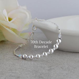 May include: A silver-toned bracelet featuring a series of round beads and small cylindrical spacers, accented with clear crystals. The text 70th Decade Bracelet is visible in the center. The design includes a metal chain extension with a clasp.