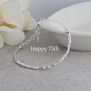 May include: A silver bracelet with a delicate design, featuring small beads and accents. The bracelet is displayed on a gray surface with the text "Happy 75th". A white flower and a white bowl are in the background.