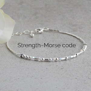 May include: Silver bracelet with small beads and a clasp. The bracelet has the word "Strength" written on it, followed by a hyphen and the words "Morse code".