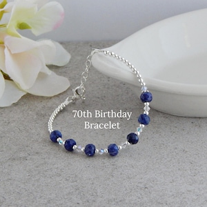 May include: A silver bracelet with dark blue beads and clear crystal accents. The bracelet is labeled "70th Birthday Bracelet" and is displayed on a gray surface next to a white dish and flowers.