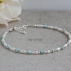 May include: A delicate bracelet featuring alternating light blue beads and white pearls, strung on a silver chain. The bracelet is displayed on a gray surface, with the word "larimar" in script. A white flower is in the background.