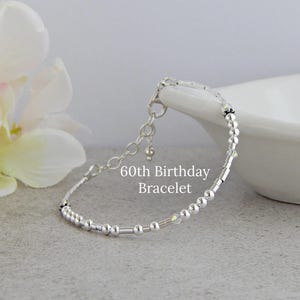 May include: A silver-toned beaded bracelet featuring a mix of small round and cylindrical beads with clear crystal accents. The bracelet includes a delicate chain extension with a small dangling bead and displays the text 60th Birthday Bracelet.
