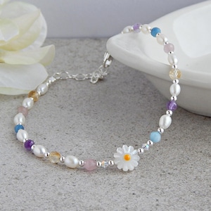May include: A delicate silver bracelet with a daisy charm and colorful gemstone beads. The bracelet features a mix of white pearls, blue, purple, pink, and yellow gemstones.