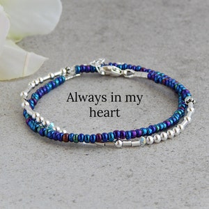 May include: A delicate bracelet with two strands of iridescent blue and silver beads. The bracelet has a silver clasp and the words "Always in my heart" are printed on a white background.