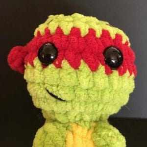 May include: A handmade, plush toy featuring a green head and body with a yellow chest. The toy has a red mask and a small red head covering. Large, black, shiny eyes and a stitched smile complete the design. The toy is likely a character from a popular franchise.