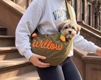 Puede incluir: Un transportín de bandolera para mascotas en color verde oliva con el nombre Willow bordado en letra naranja, decorado con pequeñas aplicaciones de zanahorias y flores, diseñado para llevar a un perro pequeño sobre el pecho.