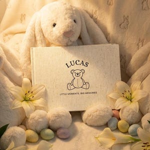 May include: A cream-coloured baby photo album with the name "LUCAS" and a teddy bear illustration. Soft white bunny toys and white lilies surround the album. Pastel-coloured Easter eggs are also present.