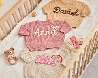 May include: Three personalized baby sweaters in a crib. One is peach with "Daniel" in brown script, another is pink with "Anna" in white script, and the third is cream with "Ellie" and a rainbow. A small bow bag and a teddy bear rattle are also present.