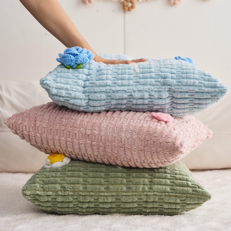 May include: A stack of three rectangular pillows with a textured, grid-like fabric surface. The top pillow is light blue with a blue crocheted flower, the middle is light pink with a small pink accent, and the bottom is sage green with a white and yellow flower.