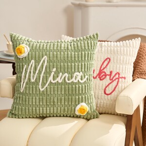 May include: Two decorative corduroy throw pillows. One is light green with the name "Mina" embroidered in white. The other is off-white with the word "by" embroidered in red. Both pillows have a floral accent.