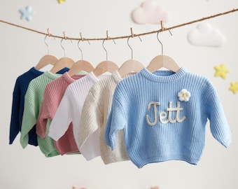 May include: A row of baby sweaters in various pastel colors, including blue, green, pink, white, and beige, hang on wooden hangers. The blue sweater has the name "Jett" embroidered on it, along with a small flower. The sweaters are displayed against a white background.
