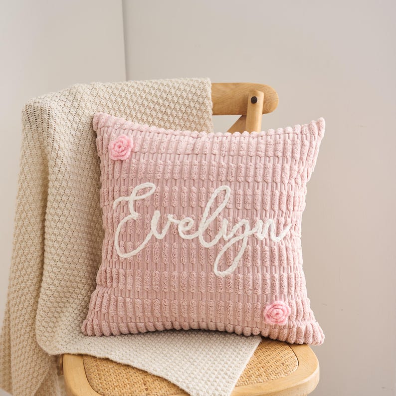 May include: A square, light pink, textured corduroy throw pillow featuring the name Evelyn embroidered in white cursive script across the center, accented with two small, light pink, three-dimensional fabric flowers in opposite corners.
