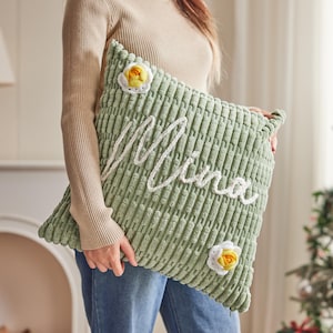 May include: A light green corduroy pillow with the name "Mina" embroidered in white. The pillow features two decorative crochet flowers with yellow centers and white petals. The pillow is approximately 18 inches square.
