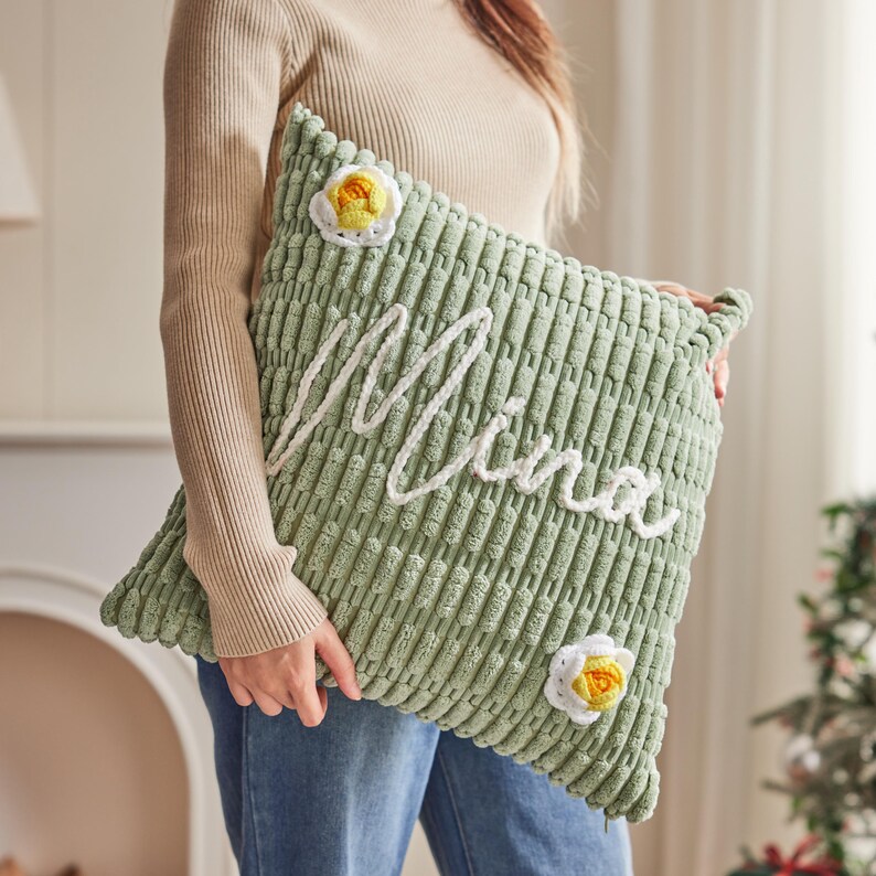 May include: A light green corduroy pillow with the name "Mina" embroidered in white. The pillow features two decorative crochet flowers with yellow centers and white petals. The pillow is approximately 18 inches square.