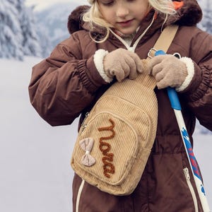 May include: A beige corduroy sling bag featuring a front zippered pocket with the name Nora embroidered in brown yarn and a small beige fabric bow attached to the side. The bag has a single adjustable shoulder strap and silver metal hardware.