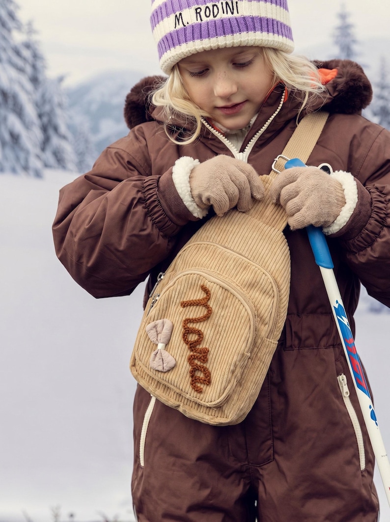 May include: A beige corduroy sling bag featuring a front zippered pocket with the name Nora embroidered in brown yarn and a small beige fabric bow attached to the side. The bag has a single adjustable shoulder strap and silver metal hardware.