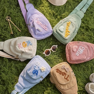 May include: Six corduroy sling bags in pastel colors including lavender, mint green, cream, pink, light blue, and tan. Each bag features a front zippered pocket with personalized embroidered names or initials and small decorative appliques.