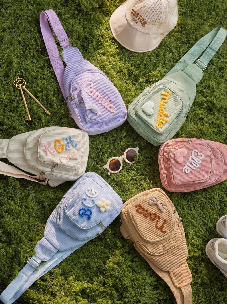 May include: Six corduroy sling bags in pastel colors including lavender, mint green, cream, pink, light blue, and tan. Each bag features a front zippered pocket with personalized embroidered names or initials and small decorative appliques.
