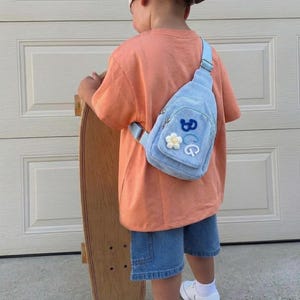 May include: A light blue corduroy sling bag featuring a small white flower patch and the blue and white embroidered letters BCG. The bag has a single adjustable strap and a zippered main compartment, worn over an orange t-shirt and denim shorts.