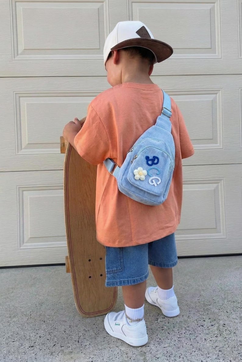 May include: A light blue corduroy sling bag featuring a small white flower patch and the blue and white embroidered letters BCG. The bag has a single adjustable strap and a zippered main compartment, worn over an orange t-shirt and denim shorts.