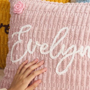 May include: A pink corduroy pillow with the name "Evelyn" embroidered in white script. A small pink crocheted flower is attached to the top left corner. The pillow has a textured surface and is part of a home decor collection.