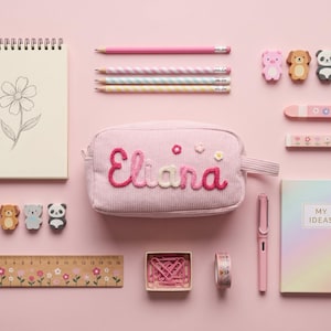 Könnte beinhalten: Ein rosafarbenes Cord-Etui mit dem eingestickten Namen Eliana in Pink und Weiß, umgeben von Schreibwaren wie einem Spiralblock mit Blumenskizze, vier gestreiften Bleistiften, acht Tierradiergummis, zwei Blumenlinealen, einer Schachtel mit rosa Büroklammern, einer Rolle Washi-Tape, einem rosa Füllfederhalter und einem pastellfarbenen Notizbuch mit der Aufschrift My Ideas.